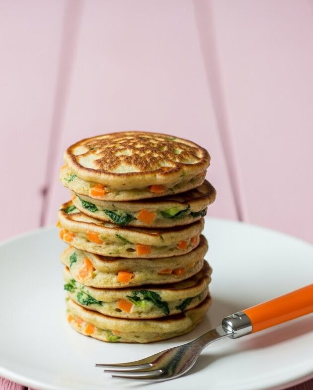 Stack of mini veggie pancakes for toddlers with visible carrots and greens on a white plate with an orange-handled fork