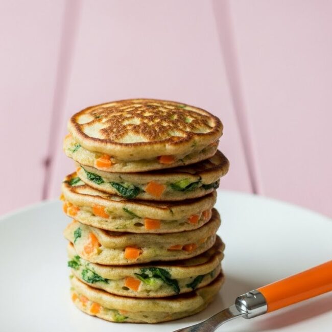 Stack of mini veggie pancakes for toddlers with visible carrots and greens on a white plate with an orange-handled fork